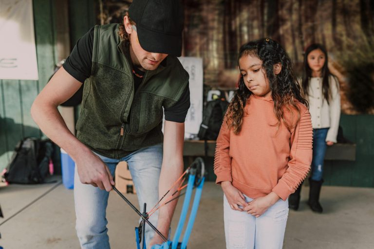 A man instructs a young girl in bow usage during an outdoor archery session.