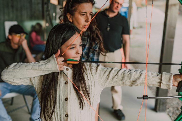 A young girl focused on archery training at an outdoor range with guidance from adults.