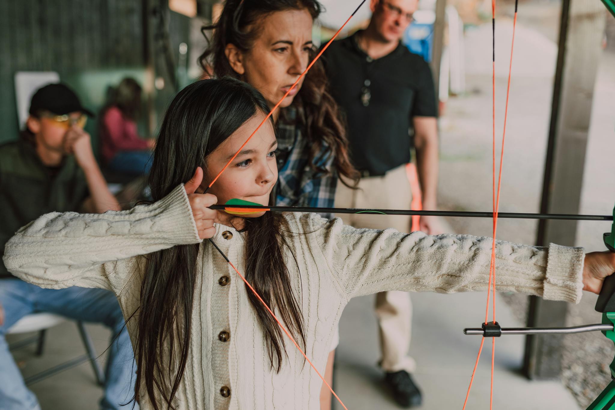 A young girl focused on archery training at an outdoor range with guidance from adults.