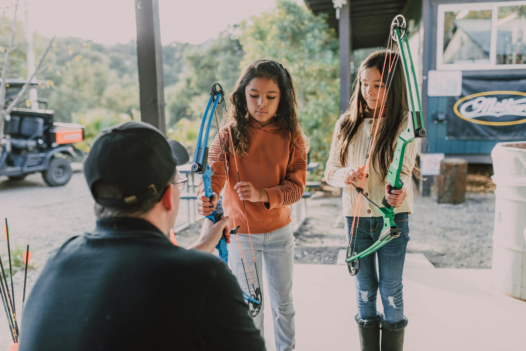 Two girls learn archery with a mentor, holding bows in an outdoor setting.