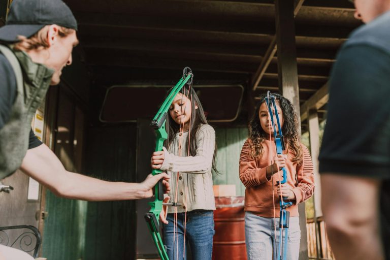 Two young girls learning archery outdoors with an instructor, holding recurve bows.